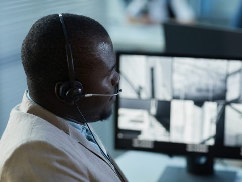 Side view portrait of black man wearing headset while watching surveillance camera feed in security and monitoring office